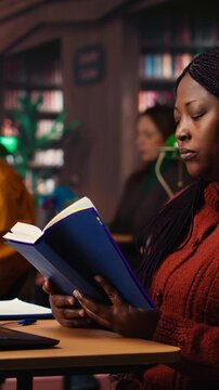 Vertical Video Black female student taking notes surrounded by books and study materials, preparing for a master dissertation and researching academic resources at the campus library. Camera B.