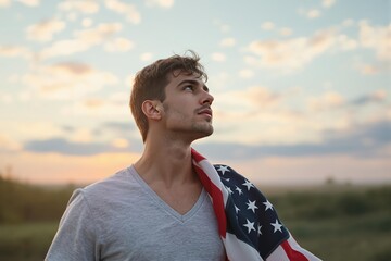 Young Caucasian man with an American flag draped over his shoulder, looking upwards with a pensive expression against a sunset sky, symbolizing patriotism and hope.