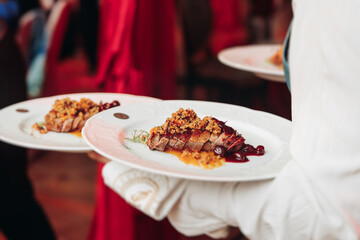 Waiter in uniform serving dishes on party event, server carrying plates with food at the table,...
