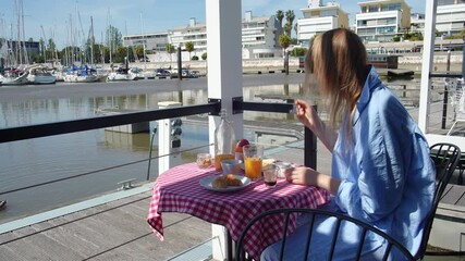 Young woman relaxing in blue bathrobe, enjoying morning breakfast at waterfront marina, sipping orange juice and savoring pastry with scenic harbor and boats surrounding her - Powered by Adobe