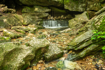 Waterfall Flowing from a Gated Cave into a Rocky Forest Stream.