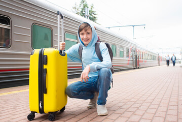 A 12 year old boy with a yellow suitcase sits squatting on the platform near the train.