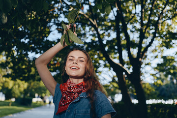 Lifestyle woman in modern boho-western denim street style smiling outdoors under green trees with...