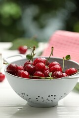 Fresh wet cherries in colander on white wooden table against blurred green background, closeup