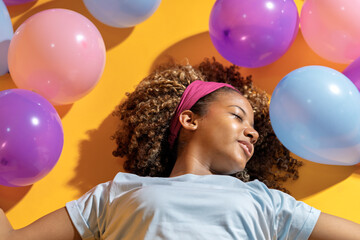Young woman lying on yellow background with colorful balloons expressing happiness