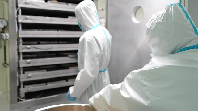 Sterile lab worker in pharmaceutical factory removes a metal container holding raw medicine or vitamin material from an industrial stainless steel chamber during preparation process.