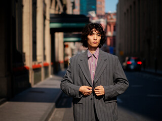 Portrait of handsome Chinese young man wearing gray suit posing in the street, young guy with black curly hair with urban background. Male fashion, cool Asian young man lifestyle.