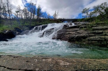 The waterfall of Rakkasjokk in Swedish Lapland flows over smooth bedrock into a turquoise pool,...