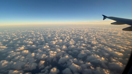 Fototapeta premium Airplane wing visible above a vast expanse of white, fluffy clouds under a clear blue sky, seen from an airplane window.