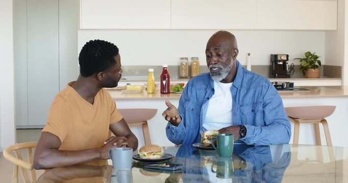 African American father and adult son leaning forward talking and eating sandwiches in home kitchen