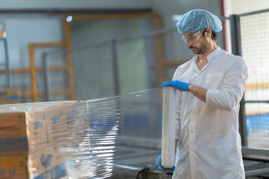 A worker in a food packaging factory wraps boxes with plastic film. He wears safety gear, including gloves, a hairnet, and protective glasses, ensuring hygiene and efficiency in production.