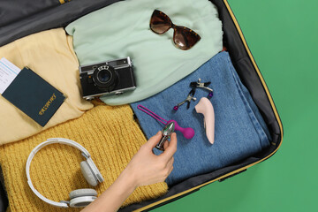 Woman putting sex toys into suitcase with clothes and accessories on green background, closeup