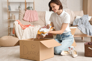 Young pretty woman folding baby clothes in donation cardboard box at home