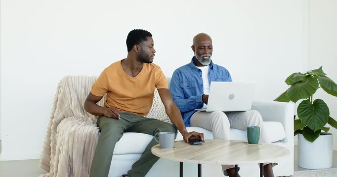 African American son teaching father sitting on sofa using smartphone and mouse on silver laptop