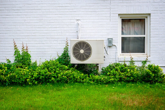 mini split air conditioner / heat pump exterior unit mouted ground floor  brick wall white of older apartment building green vegetation shot toronto beaches in summer room for 