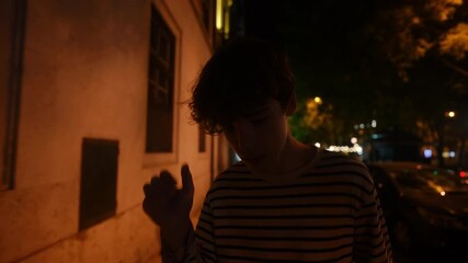 Teenage boy walking alone through dimly lit urban street at night, passing parked cars and shadowy buildings under soft street lamp illumination, embodying quiet contemplation