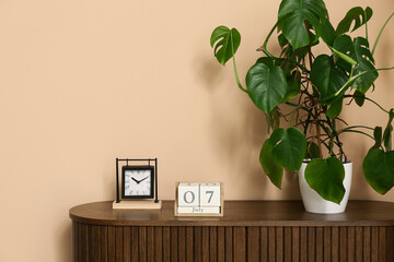 Cube calendar with date 7 JULY, clock and palm plant on chest of drawers near beige wall