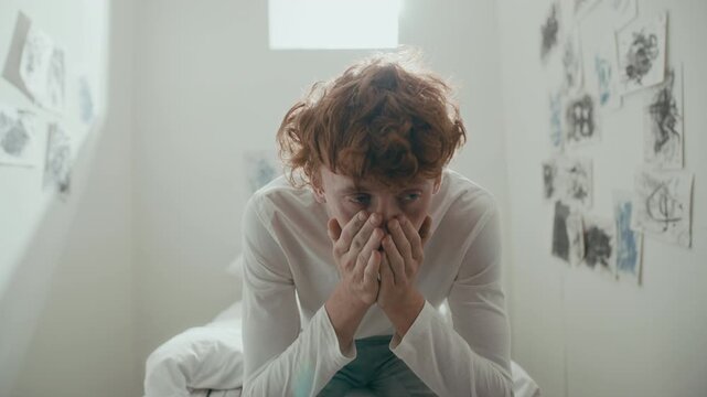 Young anxious male patient leaning forward with head in hands, then staring away in emotional distress while sitting on bed in psychiatric ward with expressive drawings on walls