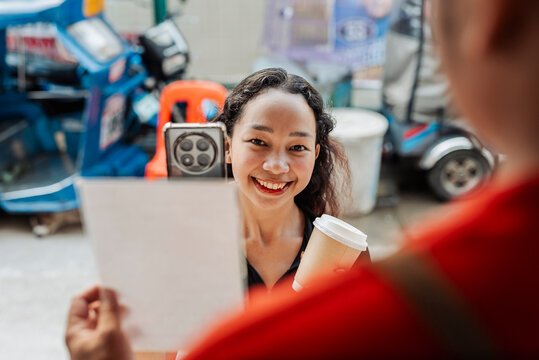 Smiling customer holds takeaway coffee cup while completing QR code payment with smartphone at a small café stand. - Powered by Adobe