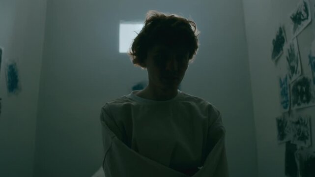 Young depressed patient in straitjacket sitting on bed in asylum room with flickering light, staring ahead with sad expression as suffering from mental disorder