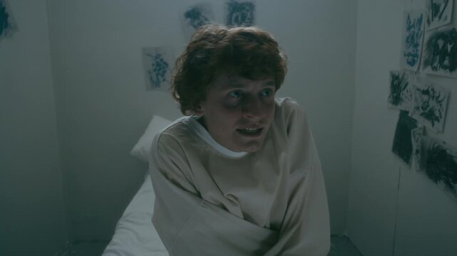 Young psychiatric patient restrained in straitjacket screaming with rage at camera, sitting alone in isolation room in asylum, surrounded by ink drawings. Video portrait