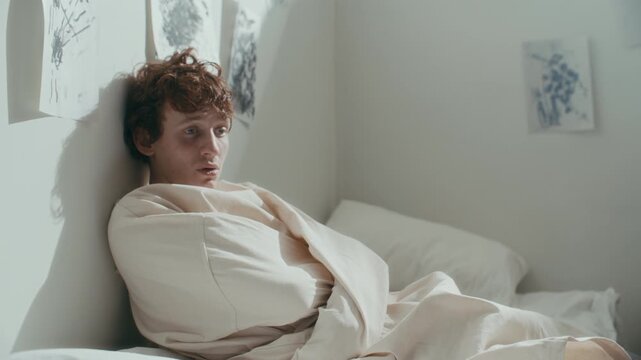 Disoriented young male patient in straitjacket leaning against wall and listening to doctor talking to him during clinical session in psychiatric ward