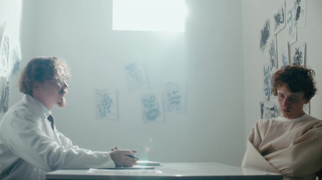 Young male patient in restraint gown sitting across from psychiatrist talking to him during mental health therapy session in sunlit hospital ward with abstract drawings on wall