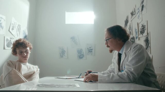 Professional doctor attentively listening to young male patient in straitjacket sitting across table in psychiatric ward with abstract drawings covering walls. Zoom-out shot