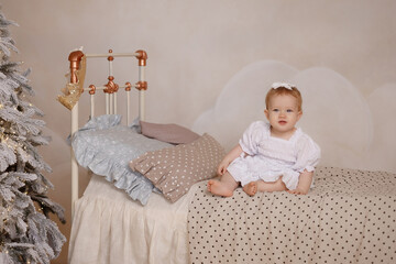 Caucasian baby girl, about 1 year old, in white dress with bow, sits on bed, front view, looks at camera. Vintage bed, polka dot bedding, white wall, Christmas tree.Concepts: childhood, holiday, home.