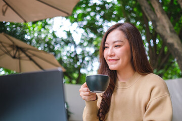 Portrait image of a young woman drinking coffee while working on laptop computer in the outdoors cafe