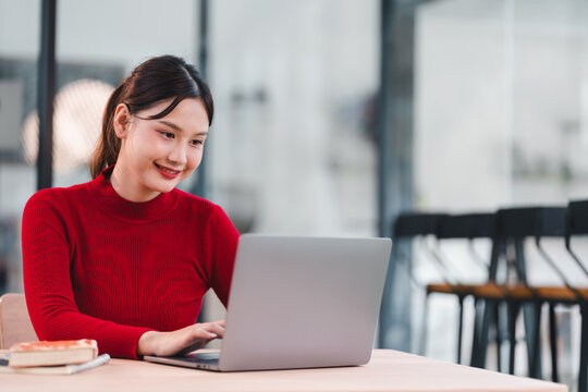 Young woman working on laptop in modern office, smiling and focused on screen, casual red sweater, bright and clean workspace - Powered by Adobe