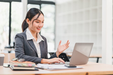 Young businesswoman working on laptop in modern office, smiling and gesturing during video call, professional atmosphere with natural light