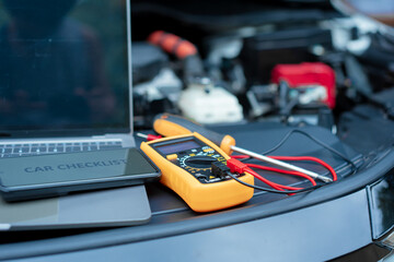 Essential car maintenance tools set up: a laptop rests on the open engine bay, a smartphone displaying “CAR CHECKLIST” on top, and a multimeter with test leads placed nearby