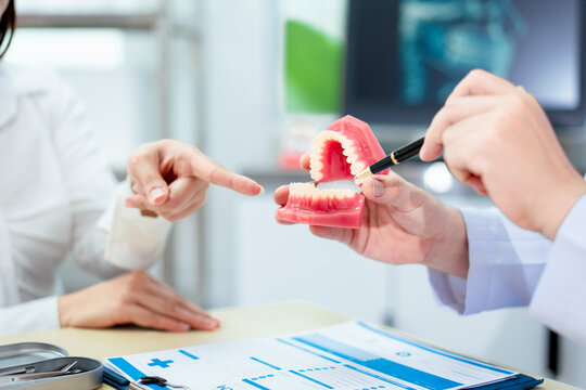 An Asian male dentist explains dental health to a middle-aged Asian female patient while holding a dental model, demonstrating oral care and treatment procedures in a clinical consultation setting