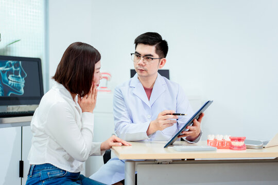 An Asian male dentist explains dental health to a middle-aged Asian female patient while holding a dental model, demonstrating oral care and treatment procedures in a clinical consultation setting
