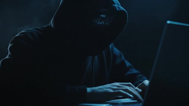 Anonymous Hacker Wearing Hoodie Mask Typing on Laptop in Dark Room with Blue Smoke and Low Light Atmosphere for Cybercrime and Data Breach Concept