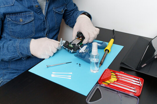 Electronic technician wearing gloves disassembling and repairing broken gaming controller using precision tools and online tutorial on tablet