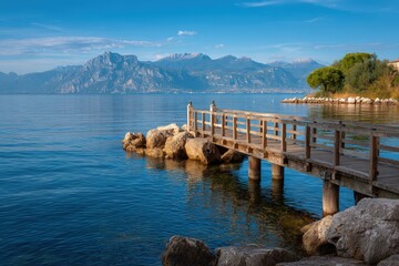 Naklejka premium Wooden pier extends into calm blue water distant mountains under clear sky