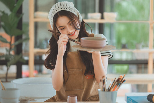 Asian woman artist painting handmade ceramic product in pottery studio – creative process of decorating sculpture with brush for small business, art workshop, and modern craft design startup