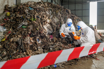 Workers in protective suits and masks inspecting large pile of waste and debris inside industrial...