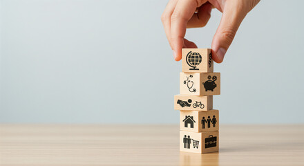 Hand carefully placing a wooden block with a globe icon atop a stack of similar blocks, each displaying various life-planning symbols.