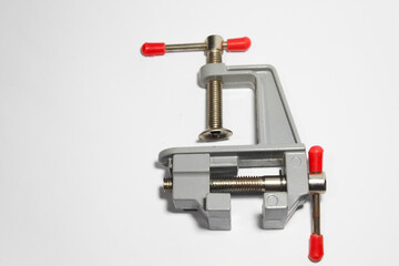A close-up image of a metal bench vise clamp with red plastic handle tips, isolated on a white background. This hand tool is used for holding objects securely in place during work or repairs.