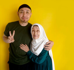 Affectionate Asian Young Muslim Man Hugging His Mother And Showing Peace Sign On Yellow Background
