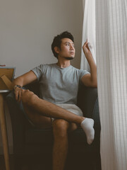 Young Thai man stretching arms while sitting relaxed on chair beside window light in a quiet minimalist Thai apartment with natural daylight