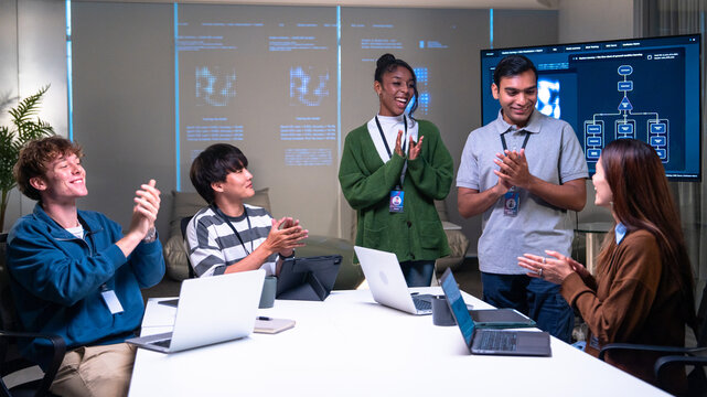 Diverse group of programmer developers in office clapping and smiling while discussing code on laptops and digital screens, showing teamwork and positive energy in modern workspace