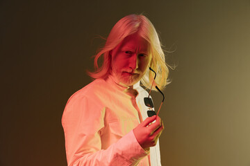 Stylish man with long white hair holding sunglasses in a dramatic pose, against a gradient...