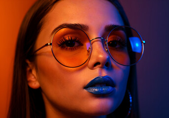 Stylish close-up of a young woman with straight brown hair, wearing round orange-tinted glasses and bold blue lipstick, captured under dramatic orange and blue lighting for a modern, edgy look.