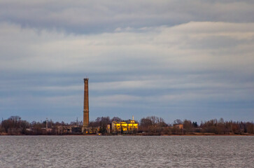 Tall industrial chimney near calm lake under cloudy evening sky view. Distant industrial chimney...