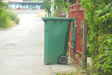 Naklejka premium A green trash bin with an open lid stands near the fence of a house - close-up against road background. Symbol of local waste control, responsible living, environmental habits and eco-lifestyle.
