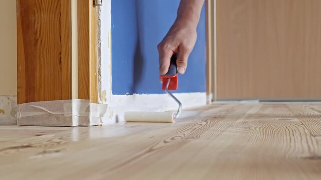 Close-up of a hand using a paint roller to apply transparent varnish to a pine wood floor near a door frame during home renovation, static camera, clear daylight, smooth motion for DIY tutorial